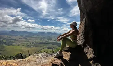 Trilheira sentada em um mirante de pedra na Pedra das Caveiras, observando a vista panorâmica das montanhas e vales de Atílio Vivacqua sob céu azul.