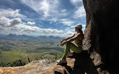 Trilheira sentada em um mirante de pedra na Pedra das Caveiras, observando a vista panorâmica das montanhas e vales de Atílio Vivacqua sob céu azul.