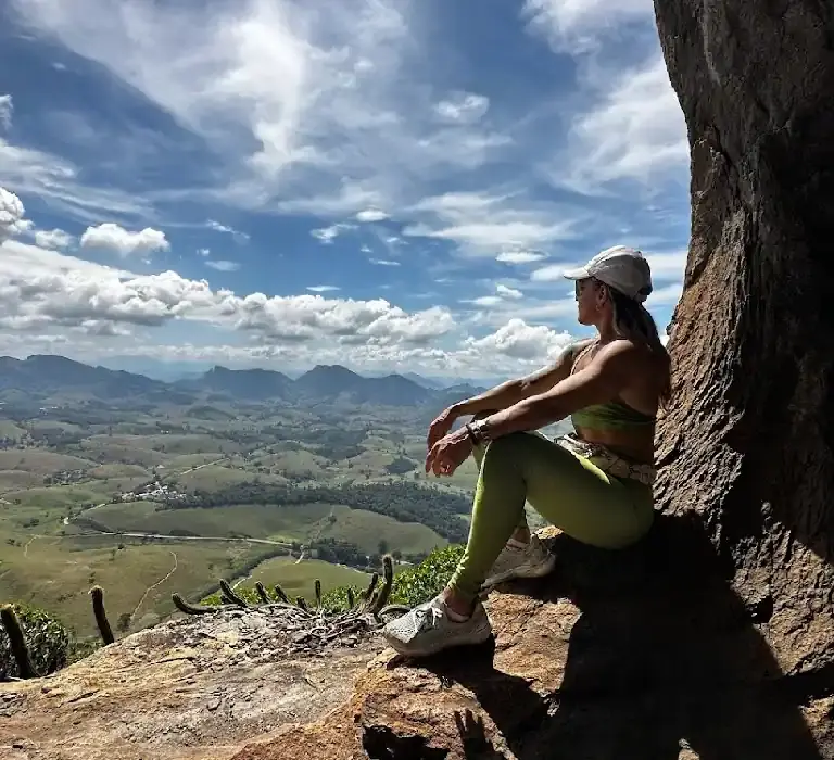 Trilheira sentada em um mirante de pedra na Pedra das Caveiras, observando a vista panorâmica das montanhas e vales de Atílio Vivacqua sob céu azul.