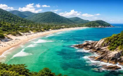 Paisagem aérea de uma praia paradisíaca no Sul do Brasil, com mar verde-esmeralda, areia branca e vegetação nativa, representando o litoral de Santa Catarina.
