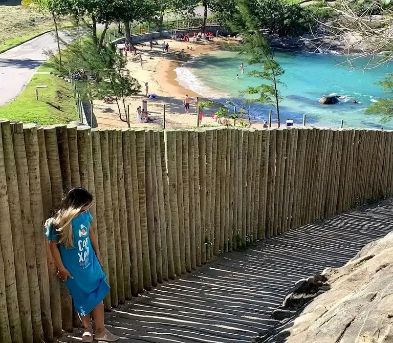 Mulher observando o mar turquesa em uma praia tranquila do Espírito Santo, com acesso por uma escadaria de madeira rústica.