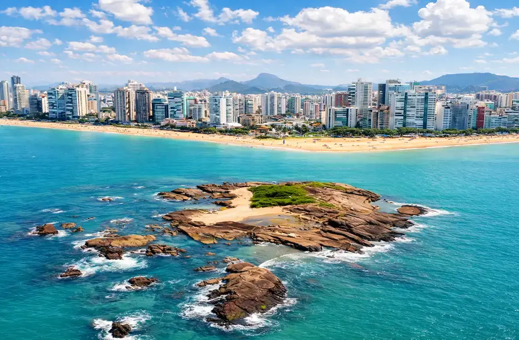 Vista panorâmica da Ilha Pituã em Vila Velha, com suas águas cristalinas e barcos de pescadores ancorados próximos às piscinas naturais, com a Praia de Itapuã ao fundo.