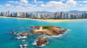 Vista panorâmica da Ilha Pituã em Vila Velha, com suas águas cristalinas e barcos de pescadores ancorados próximos às piscinas naturais, com a Praia de Itapuã ao fundo.
