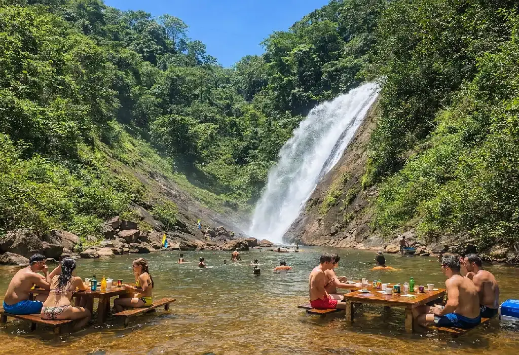 Turistas relaxando em mesas de madeira dentro da água em uma cachoeira cercada por mata verde.