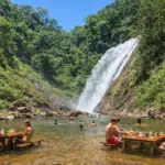 Turistas relaxando em mesas de madeira dentro da água em uma cachoeira cercada por mata verde.