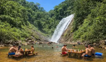 Turistas relaxando em mesas de madeira dentro da água em uma cachoeira cercada por mata verde.