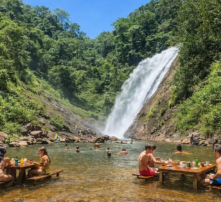 Turistas relaxando em mesas de madeira dentro da água em uma cachoeira cercada por mata verde.