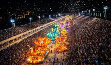 Vista panorâmica do Sambódromo lotado durante desfile de escola de samba, com carros alegóricos iluminados e arquibancadas cheias." Título da Imagem: "Carnaval 2026: A alta procura por ingressos e camarotes.