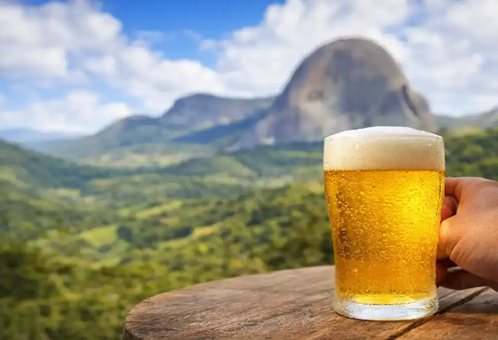 Mão segurando um copo de cerveja artesanal gelada com espuma sobre uma mesa de madeira, tendo a Pedra Azul e montanhas verdes ao fundo.