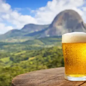 Mão segurando um copo de cerveja artesanal gelada com espuma sobre uma mesa de madeira, tendo a Pedra Azul e montanhas verdes ao fundo.