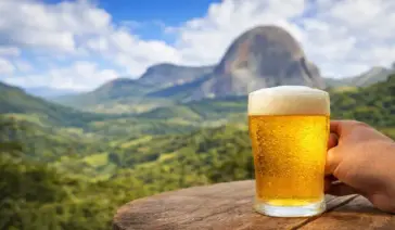 Mão segurando um copo de cerveja artesanal gelada com espuma sobre uma mesa de madeira, tendo a Pedra Azul e montanhas verdes ao fundo.