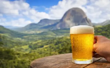 Mão segurando um copo de cerveja artesanal gelada com espuma sobre uma mesa de madeira, tendo a Pedra Azul e montanhas verdes ao fundo.