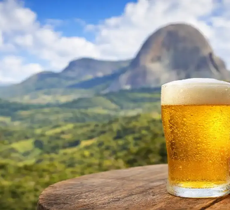 Mão segurando um copo de cerveja artesanal gelada com espuma sobre uma mesa de madeira, tendo a Pedra Azul e montanhas verdes ao fundo.