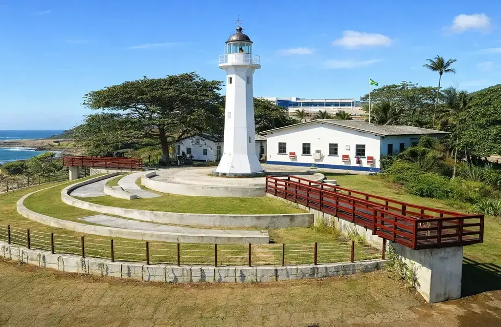 Farol Santa Luzia em Vila Velha, com sua estrutura metálica vermelha, sob um céu azul ensolarado e mar ao fundo.