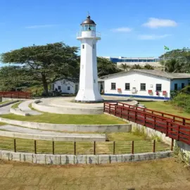 Farol Santa Luzia em Vila Velha, com sua estrutura metálica vermelha, sob um céu azul ensolarado e mar ao fundo.