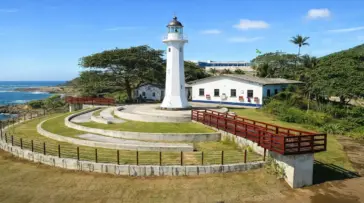 Farol Santa Luzia em Vila Velha, com sua estrutura metálica vermelha, sob um céu azul ensolarado e mar ao fundo.