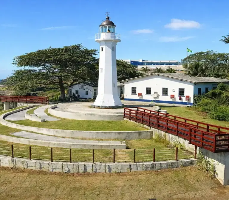 Farol Santa Luzia em Vila Velha, com sua estrutura metálica vermelha, sob um céu azul ensolarado e mar ao fundo.