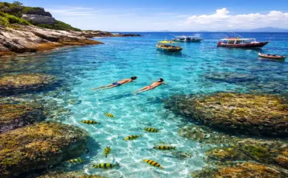 Mergulho em águas transparentes cor turquesa com peixes coloridos em primeiro plano e barcos ao fundo, representando as ilhas do Espírito Santo.
