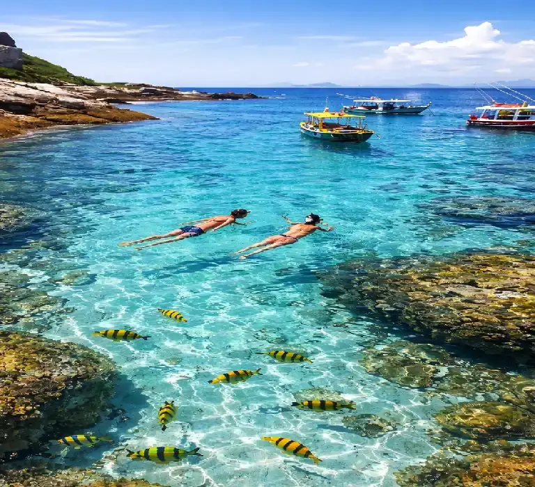 Mergulho em águas transparentes cor turquesa com peixes coloridos em primeiro plano e barcos ao fundo, representando as ilhas do Espírito Santo.
