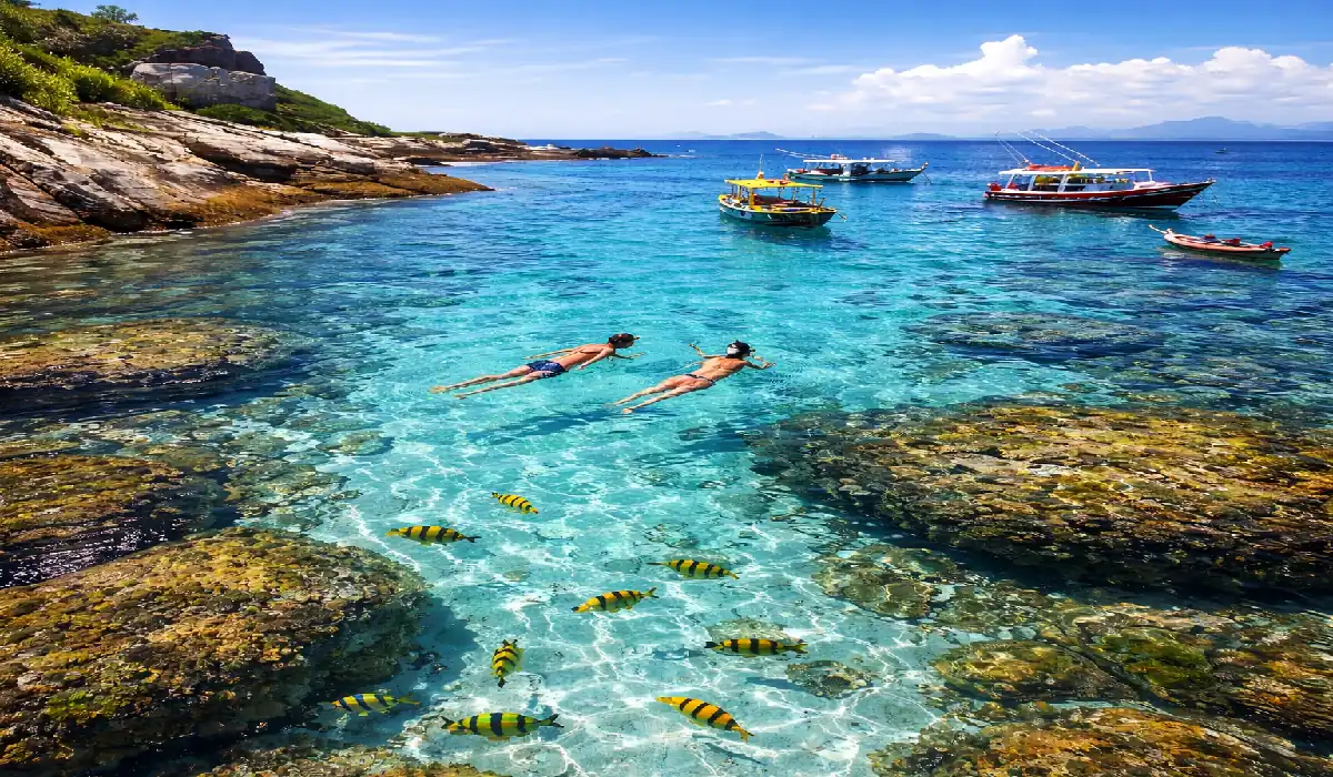 Parece Cancún, mas é no ES: 3 ilhas de água "neon" para visitar de barco neste verão. 14 Mergulho em águas transparentes cor turquesa com peixes coloridos em primeiro plano e barcos ao fundo, representando as ilhas do Espírito Santo.