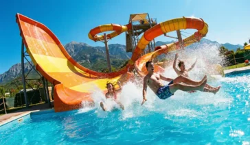 Jovens descendo em alta velocidade por um toboágua laranja e caindo na piscina, com muita água espirrando, ilustrando a diversão nos parques aquáticos