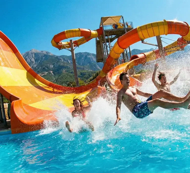 Jovens descendo em alta velocidade por um toboágua laranja e caindo na piscina, com muita água espirrando, ilustrando a diversão nos parques aquáticos