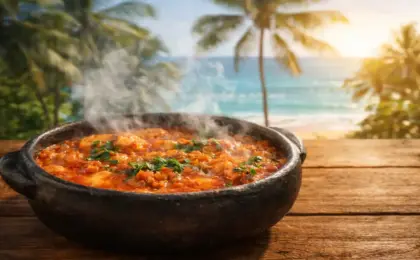 Moqueca capixaba fervendo na panela de barro preta, soltando fumaça, sobre uma mesa de madeira com vista para o mar e coqueiros ao fundo.