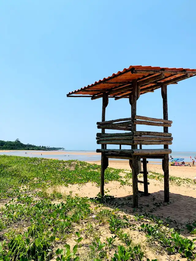 Torre de madeira na areia da Praia de Coqueiral, com vegetação e mar calmo ao fundo