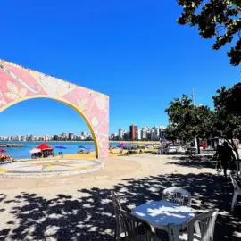 Vista aérea da Arena de Verão na Praia da Costa, Vila Velha, com quadras de esporte lotadas, palco de shows e área gastronômica na areia.