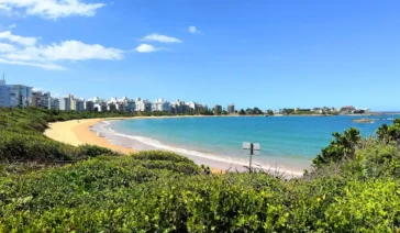 Praia urbana com mar calmo e azul-turquesa, faixa de areia clara em formato de enseada, vegetação verde em primeiro plano e prédios residenciais ao fundo sob céu azul.