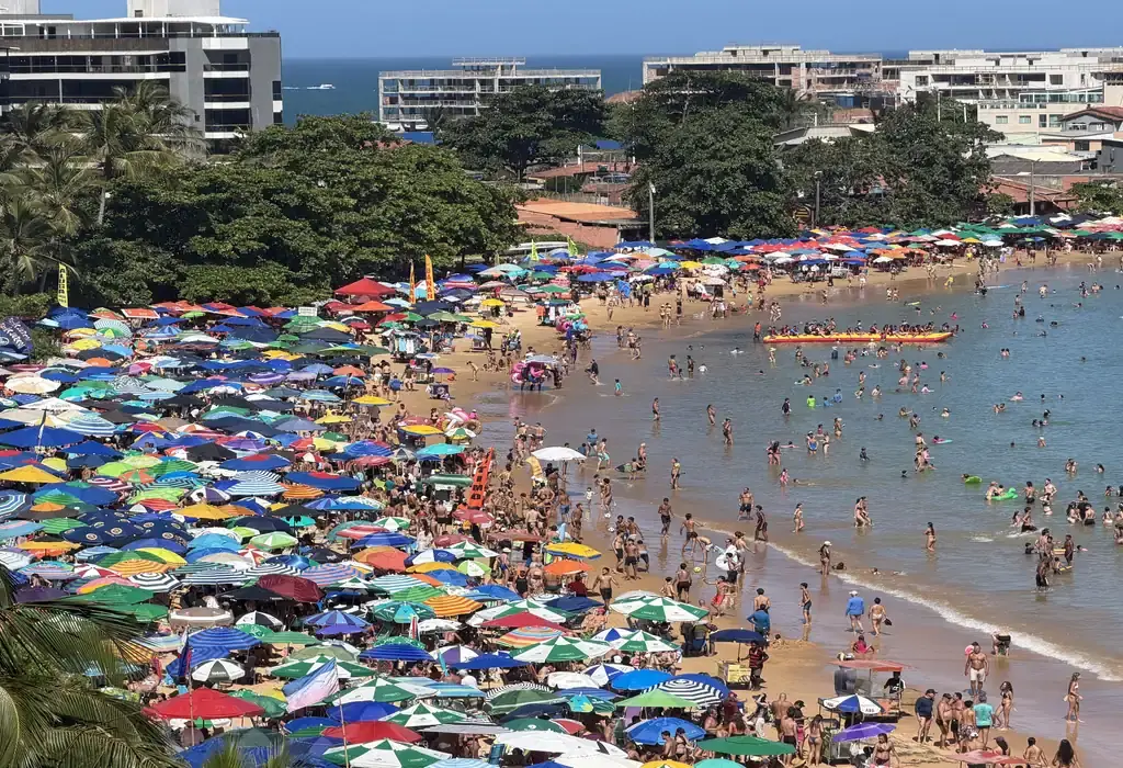 Vista panorâmica da Praia de Peracanga, em Guarapari, completamente lotada. A faixa de areia está coberta por centenas de guarda-sóis coloridos e muitos banhistas aproveitam o mar calmo da Enseada Azul em um dia de sol.