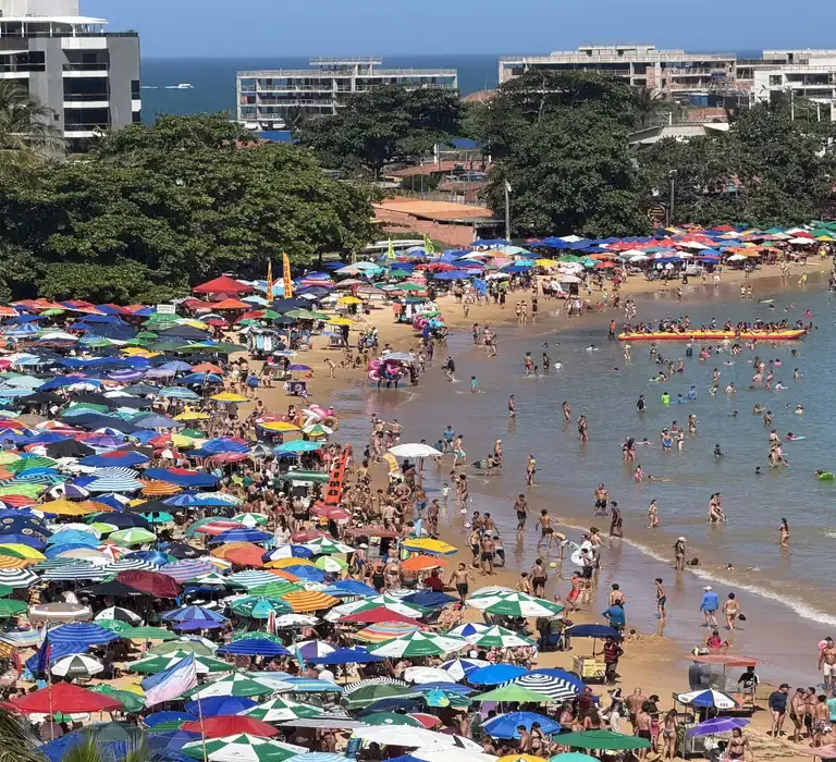 Vista panorâmica da Praia de Peracanga, em Guarapari, completamente lotada. A faixa de areia está coberta por centenas de guarda-sóis coloridos e muitos banhistas aproveitam o mar calmo da Enseada Azul em um dia de sol.