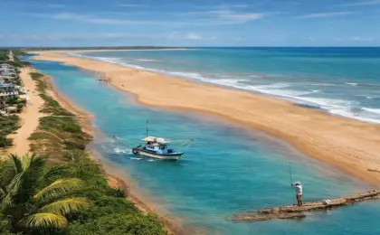 Vista aérea ensolarada da Praia de Urussuquara, no Espírito Santo, mostrando o rio de águas turquesas correndo paralelo ao mar, com um barco de pesca e vegetação nativa.