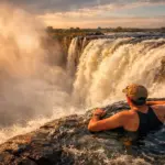 Turista no Devil’s Pool observando a Victoria Falls com arco-íris e névoa ao pôr do sol