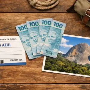 Composição fotográfica sobre uma mesa de madeira contendo uma passagem de ônibus para Pedra Azul, três notas de 100 reais brasileiras e uma foto da Pedra do Lagarto. Representação visual de um roteiro de viagem econômico nas montanhas capixabas.