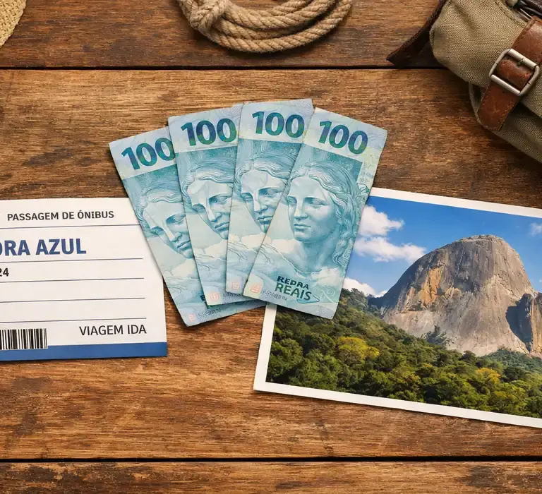 Composição fotográfica sobre uma mesa de madeira contendo uma passagem de ônibus para Pedra Azul, três notas de 100 reais brasileiras e uma foto da Pedra do Lagarto. Representação visual de um roteiro de viagem econômico nas montanhas capixabas.