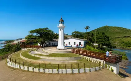 Farol Santa Luzia em Vila Velha, com sua estrutura metálica vermelha, sob um céu azul ensolarado e mar ao fundo.